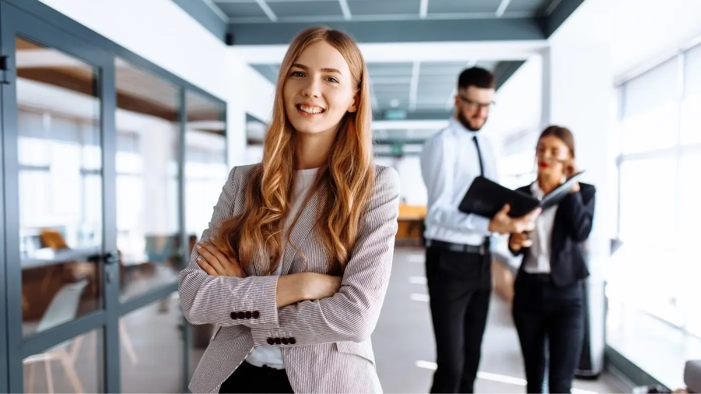 Une jeune femme dans un bureau, les bras croisés, rit avec assurance face à la caméra.